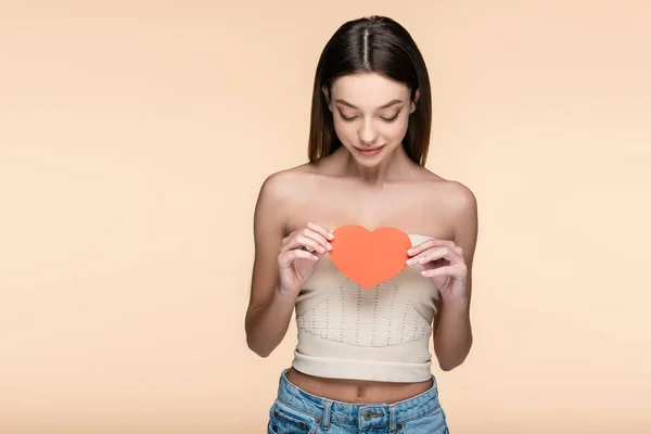 Brunette woman in crop top with bare shoulders looking at red paper heart isolated on beige — Stock Photo