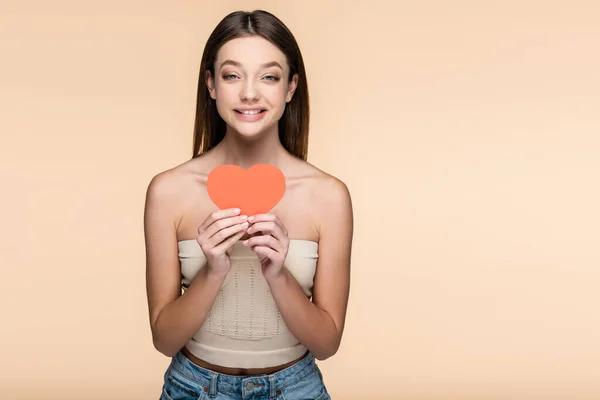 Felice giovane donna in crop top tenendo cuore di carta rossa isolato su beige — Foto stock