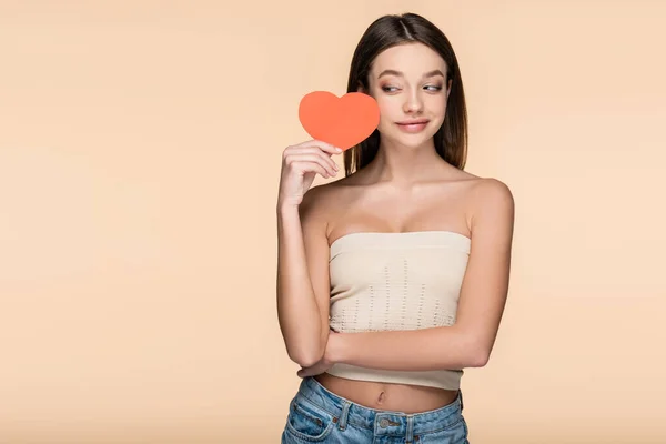 Joyful woman in crop top with bare shoulders holding red paper heart isolated on beige — Stockfoto