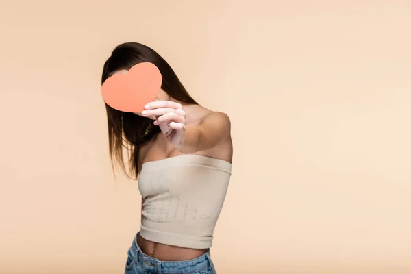 Brunette jeune femme en haut de culture avec épaules nues couvrant le visage avec du papier rouge coeur isolé sur beige — Photo de stock