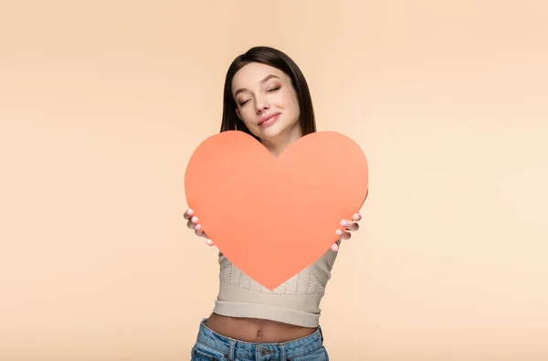 Mujer alegre con los ojos cerrados sosteniendo corazón de papel rojo aislado en beige - foto de stock