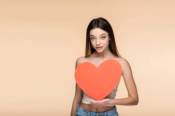 Young brunette woman with bare shoulders holding red paper heart isolated on beige — стоковое фото