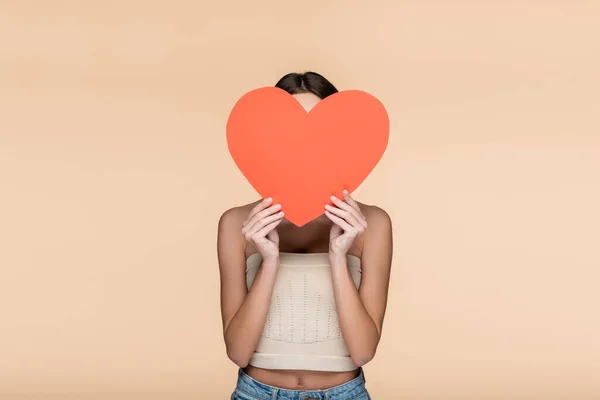 Young woman in crop top with bare shoulders covering face with red paper heart isolated on beige — стоковое фото