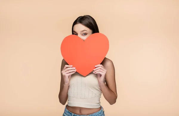 Young woman in crop top covering face with red paper heart isolated on beige — Stock Photo