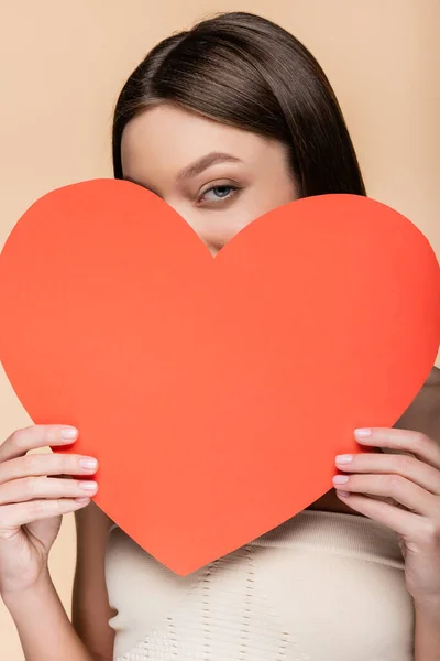 Mujer joven cubriendo la cara con papel rojo corazón cortado aislado en beige - foto de stock