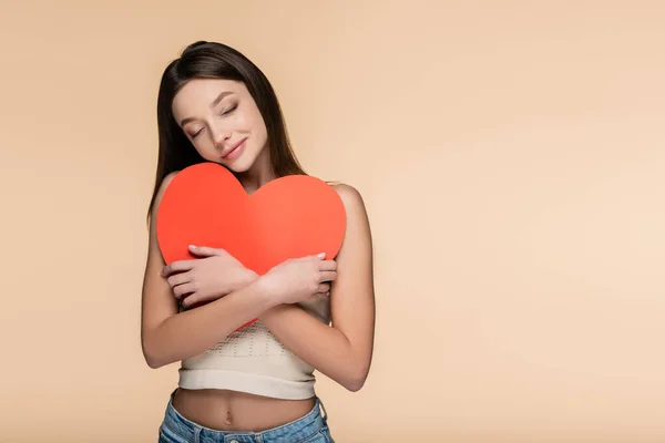 Mujer alegre con los ojos cerrados abrazando corazón de papel rojo aislado en beige - foto de stock