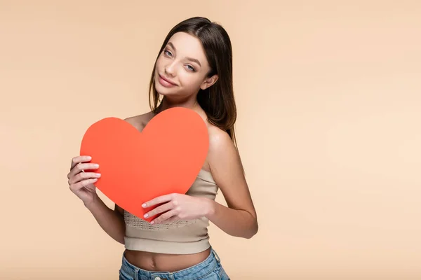 Mujer joven complacida sosteniendo corazón de papel rojo aislado en beige - foto de stock