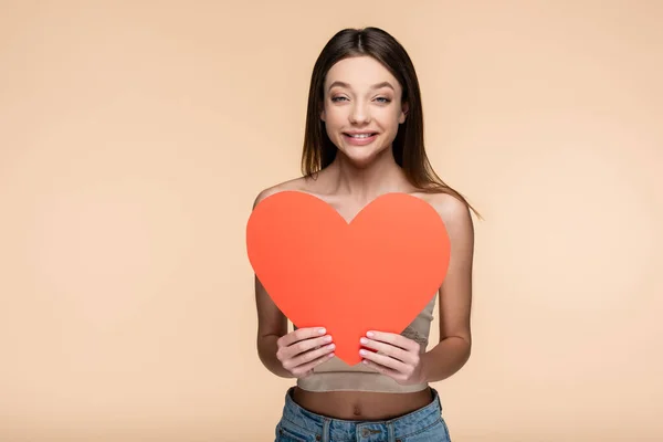 Positive young woman in crop top holding red paper heart isolated on beige — стоковое фото
