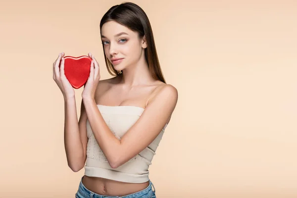 Brunette young woman holding heart-shaped metallic box isolated on beige — стоковое фото