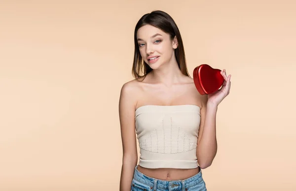 Позитивна молода жінка тримає червону металеву коробку у формі серця ізольовано на бежевому — стокове фото