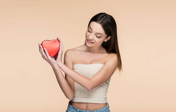 Happy young woman looking red heart-shaped metallic box isolated on beige — Stockfoto