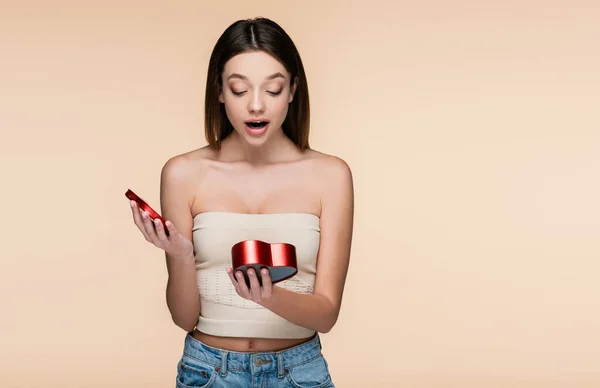 Surprised young woman looking at heart-shaped red box isolated on beige — стоковое фото