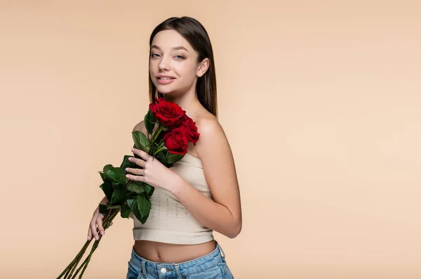 Happy young woman in crop top holding red roses isolated on beige — стоковое фото