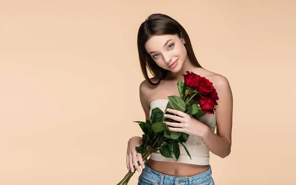 Jovem mulher satisfeita na colheita superior segurando rosas vermelhas isoladas no bege — Fotografia de Stock