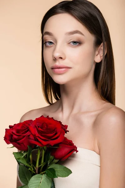 Morena joven con hombros desnudos sosteniendo rosas rojas aisladas en beige - foto de stock