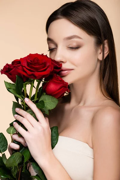 Morena joven con los ojos cerrados oliendo rosas rojas aisladas en beige - foto de stock