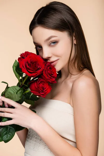 Heureuse jeune femme aux épaules nues sentant les roses rouges isolées sur beige — Photo de stock