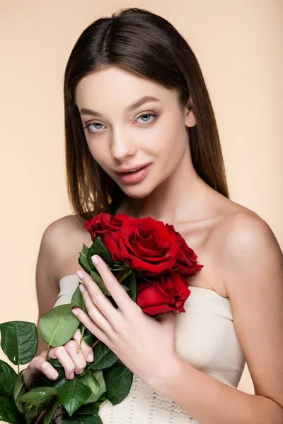 Mujer joven con hombros desnudos sosteniendo ramo de rosas rojas aisladas en beige - foto de stock