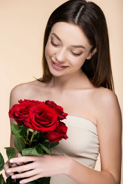 Happy woman with bare shoulders looking at bouquet of red roses isolated on beige — Fotografia de Stock