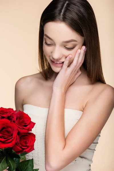 Cheerful woman with bare shoulders looking at bouquet of red roses isolated on beige — Stock Photo