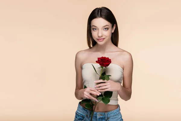 Pleased young woman with bare shoulders holding red rose isolated on beige — Stockfoto