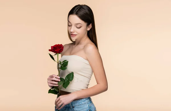 Брюнетка молодая женщина с голыми плечами, глядя на красную розу изолированы на бежевый — стоковое фото