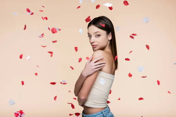 Young woman in crop top near falling rose petals on beige — стоковое фото