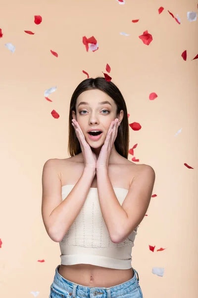 Excited young woman in crop top near falling rose petals on beige — стоковое фото