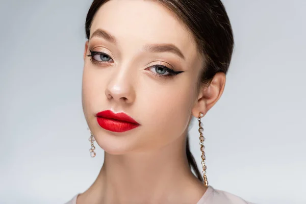 Hübsche junge Frau mit Ohrringen und roten Lippen, die vereinzelt in die Kamera auf grau blickt — Stockfoto