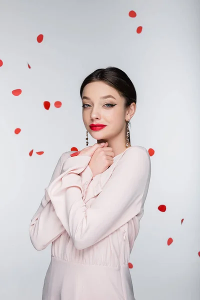 Mujer joven y complacida con maquillaje brillante cerca de caer pétalos de rosa en gris - foto de stock