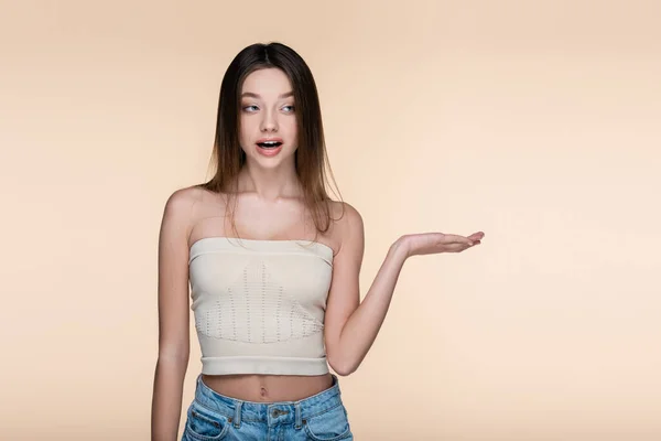 Young and shocked woman in crop top pointing with hand isolated on beige — стоковое фото