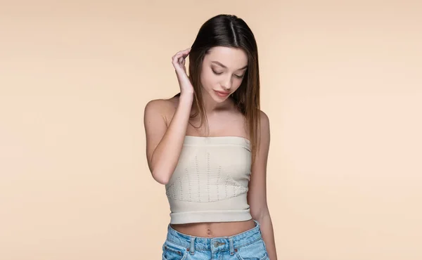 Young and shy woman in crop top adjusting hair isolated on beige — Stockfoto