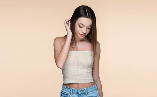 Mujer joven y tímida en la parte superior de la cosecha ajustando el cabello aislado en beige - foto de stock