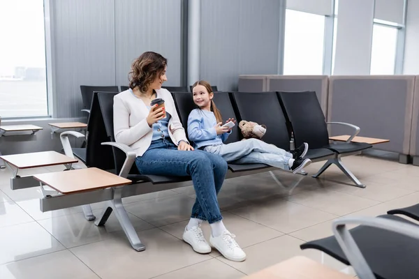 Fröhliches Kind hält Smartphone in der Hand und blickt Mutter mit Pappbecher in der Lounge des Flughafens an — Stockfoto