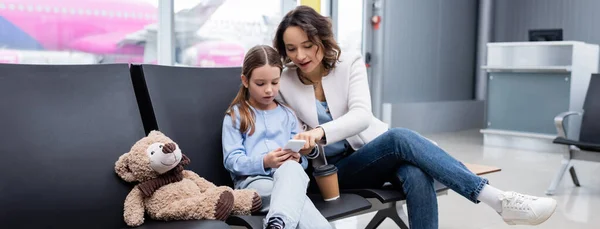 Mother with paper cup and daughter looking at smartphone in lounge hall of airport, banner - foto de stock