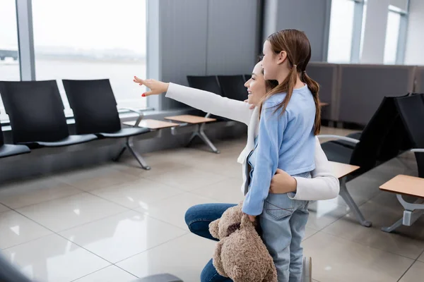 Glückliches Mädchen mit Stofftier in der Nähe einer fröhlichen Mutter, die mit der Hand auf den Flughafen zeigt — Stockfoto