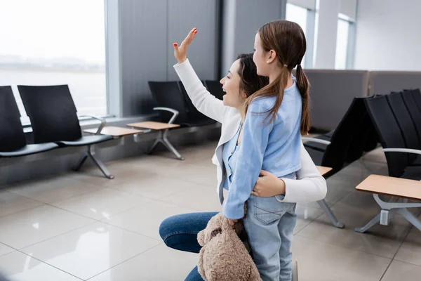 Feliz chica sosteniendo suave juguete cerca excitado madre saludando mano en aeropuerto - foto de stock