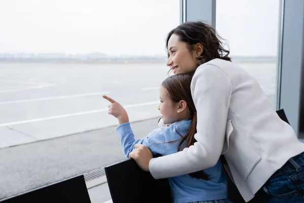 Happy kid pointing with finger at window near cheerful mother in airport — Stock Photo