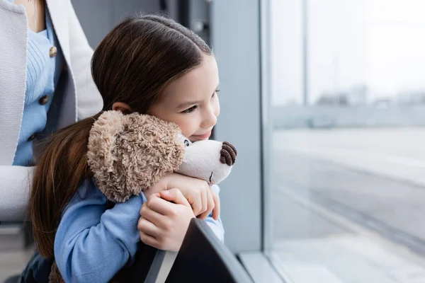 Zufriedenes Kind hält Teddybär in der Nähe der Mutter und blickt auf Flughafenfenster — Stockfoto