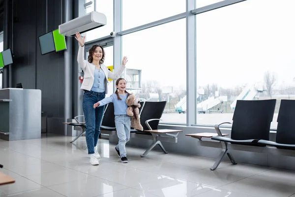 Happy mother and daughter waving hands while walking in airport lounge hall — Stock Photo