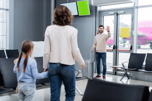Hombre feliz con el equipaje agitando la mano mientras mira a la esposa y la hija en el aeropuerto - foto de stock