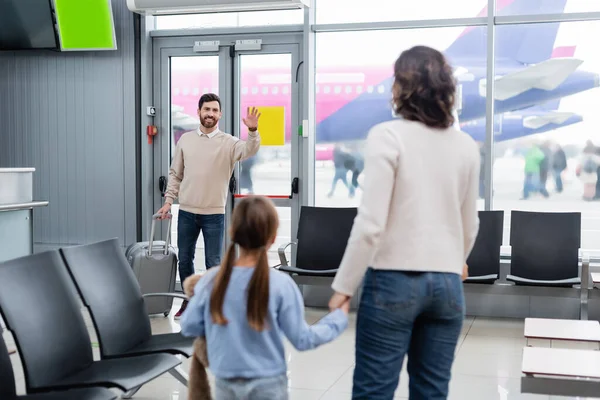 Alegre hombre con equipaje agitando la mano mientras mira a la esposa y la hija en el aeropuerto - foto de stock