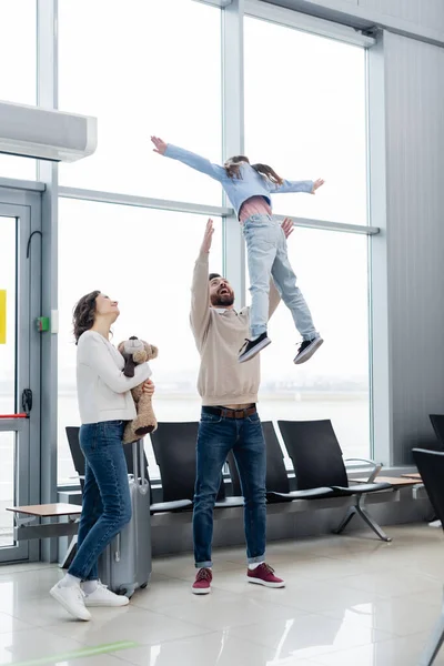 Веселий батько піднімає дочку біля дружини в залі для аеропортів — стокове фото