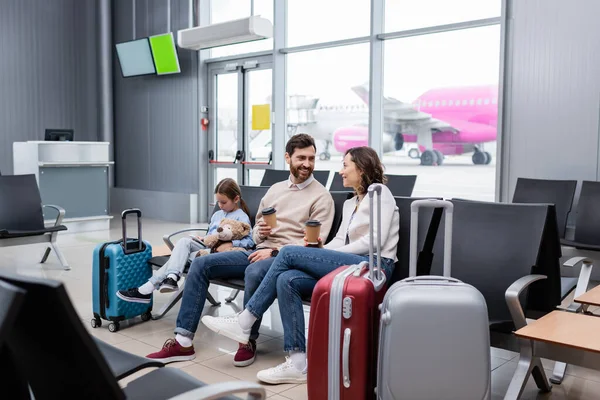 Padres felices con vasos de papel cerca de la hija y el equipaje en la sala del aeropuerto - foto de stock