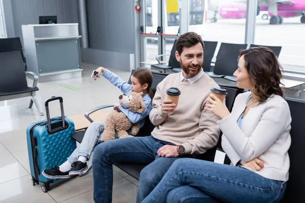 Niño tomando selfie cerca de padres alegres con vasos de papel y equipaje en el salón del aeropuerto - foto de stock