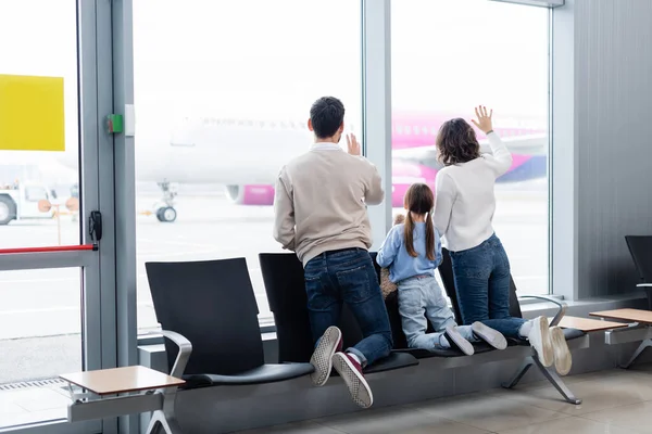 Back view of family waving hands while looking at plane through window in airport — Stock Photo