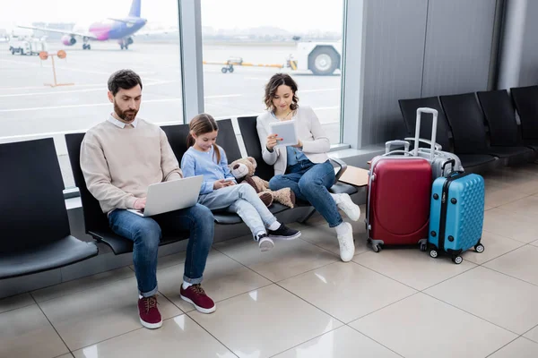 Family using devices while sitting near luggage in airport lounge — стоковое фото