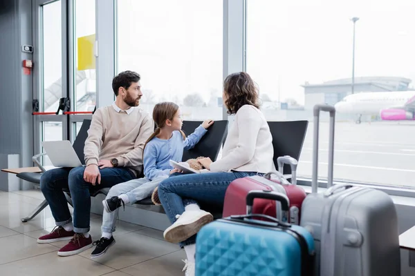Eltern halten Geräte in der Hand, während sie in der Nähe ihrer Tochter sitzen und durch das Fenster in der Flughafenlounge auf das Flugzeug schauen — Stockfoto
