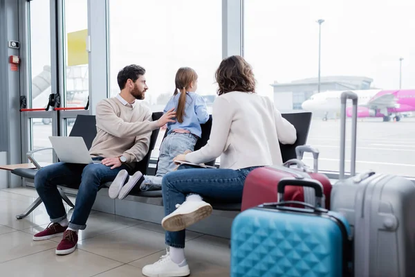 Parents holding gadgets while sitting near daughter and looking at plane through window in airport lounge — Photo de stock