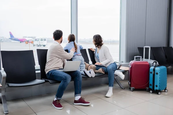 Hombre y mujer sosteniendo gadgets mientras está sentado cerca de la hija y mirando el avión a través de la ventana en el salón del aeropuerto - foto de stock